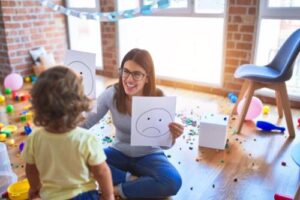 a specialist holds up a smiley face and unhappy face pictures in each hand while smiling and looking at a toddler in an applied behavior analysis program