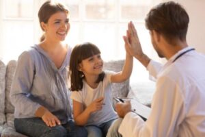 a mother sits with her young daughter who is high fiving their specialist in their parent-mediated aba therapy