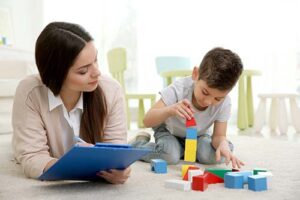 an ABA specialist sits on the floor with a young boy playing with blocks while she makes notes on her clipboard during the boy's behavior intervention and behavior support plans