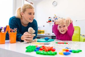 a young parent sits with her daughter at a table and watches her daughter play with toys and the parent understands why caregiver education and training is important to her child's well being