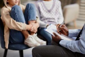a child sits in a chair with her legs up and holding onto them with her arms and her parent sitting next to her and therapist sitting across taking notes in the childs verbal behavior therapy